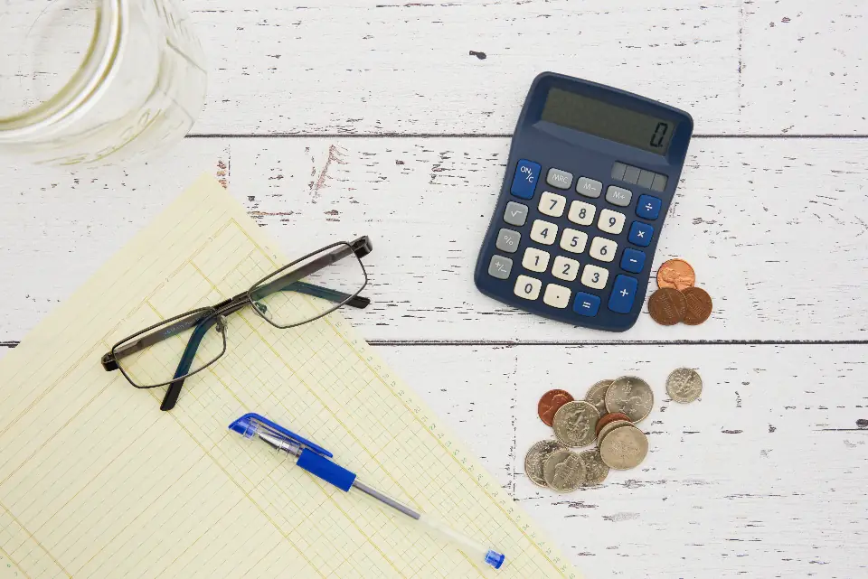 A calculator, loose coins, reading glasses and a ledger pad on a desk, ready for month-end salaries
