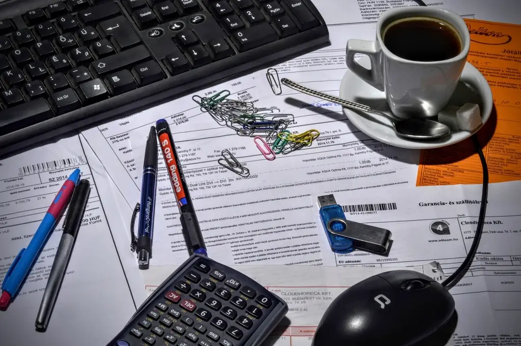 A desk crowded with papers, pens, a calculator and a coffee, the paperwork around a spreadsheet