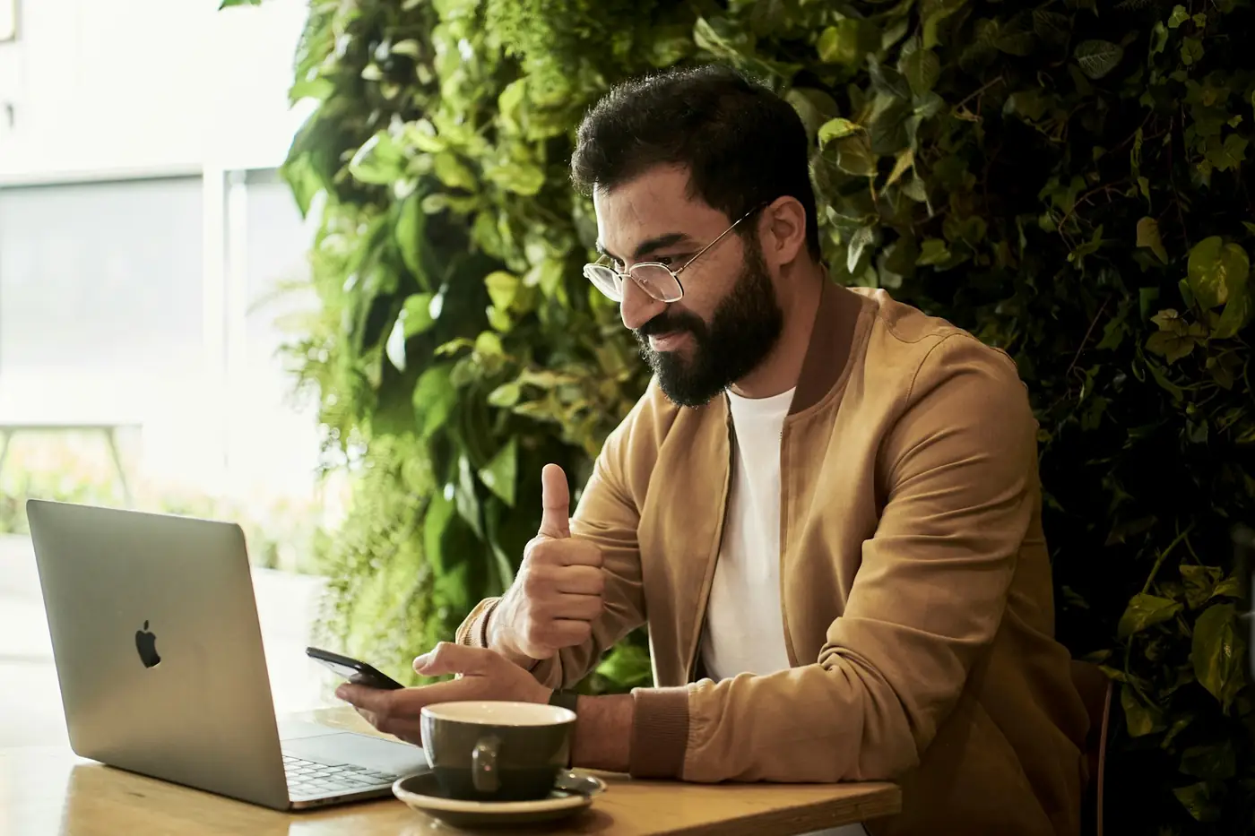 A man working on a laptop and phone at a cafe table, the way a busy client opens an approval link
