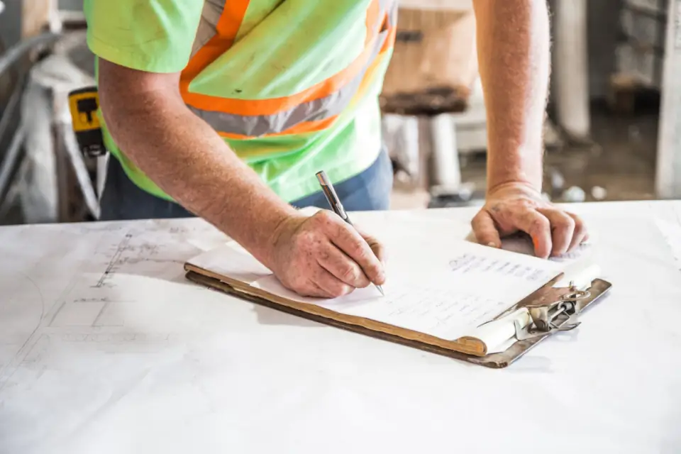 A worker in a high-visibility vest writing on a clipboard over drawings, recording the day on site